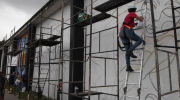 mulher está desenhando grafiti em muro. Há vários andaimes junto ao muro. #paratodosverem