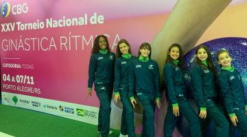 Seis meninas posam para foto na frente de outdoor do evento #paratodosverem