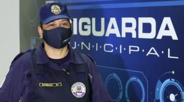 Guarda municipal uniformizada e máscara em frente a painel com o nome da corporação. #pracegover