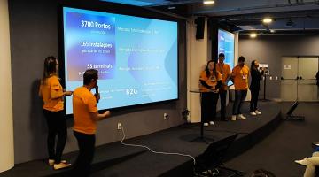 Cinco jovens estão em um palco apresentando um trabalho. Ao fundo um telão reproduz mensagens. #paratodosverem 