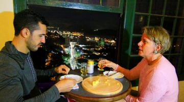Homem e mulher comem e bebem ao lado de janela com vista de Santos a noite #paratodosverem