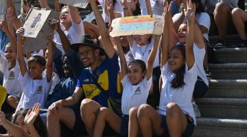 preo e criaças seguram desenhos nas mãos no alto da cabeça. #paratodosverem 