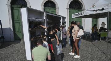 público em frente a estante de livros ao ar livre. #paratodosverem