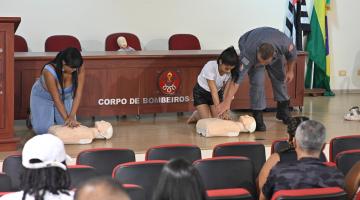 duas mulheres simulam massagem cardíaca em bonecos. militar dos bombeiros está ao lado de uma delas orientando. #paratodosverem
