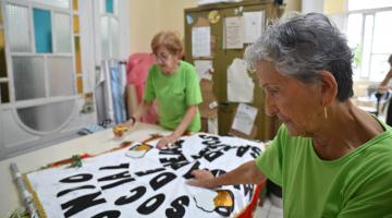 duas mulheres fazem costura em tecido com texto escrito. #paratodosverem
