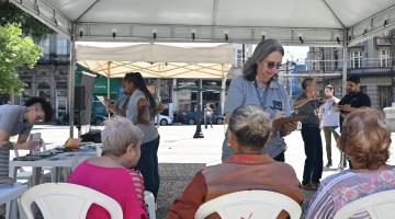 pessoas sentadas sob tenda armada em praça. #paratodosverem 