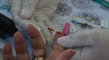 mãos com luvas usam objeto cortante para furar dedo de uma pessoa. Gota de sangue é colhida para exame. #paratodosverem 