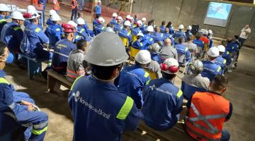 auditório cheio de operários de costas para a foto. Eles estão atentos a um homem que fala ao fundo, ao lado de um telão. Todos os operários usam uniforme e capacete. #paratodosverem