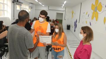 Dois voluntários usando camiseta de identificação recebem certificado de um homem que está de costas. A vice-prefeita está em pé, à direita. #paratodosverem