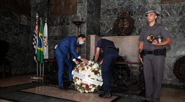 Dois homem depositam coroa de flores junto a mausoléu. #Paratodosverem