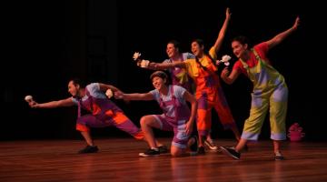 Artistas estão em palco de teatro, todos vestidos de palhaço e com flores nas mãos estendidas para a frente. #paratodosverem