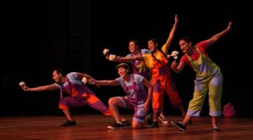 Artistas estão em coreografia no palco. Eles seguram flores nas mãos. #Paratodosverem