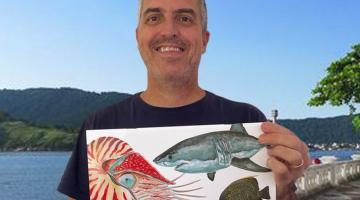 o artista plástico sorri para a foto segurando o livro-pôster com as duas mãos. Ele está à frente das muretas da praia com o mar ao fundo. #paratodosverem