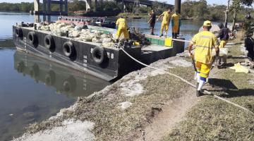 homens uniformizados colocam sacos de lixo em barcaça sobre a água. Ao fundo há uma ponte. Eles estão pisando num gramado sobre um pequeno cais à beira d'água. #paratodosverem