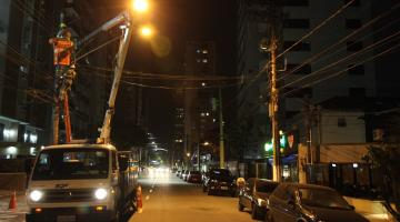 caminhão com braço basculante e cesto no topo está parado em rua para manutenção de iluminação no alto de um poste. #paratodosverem