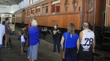 pessoas estão paradas ouvindo orientações de guia diante de antigo carro ferroviário. #paratodosverem 