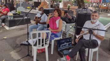 dois homens tocando instrumentos e uma mulher ao centro cantando, todos sob uma tenda, com campas ao fundo. #paratodosverem