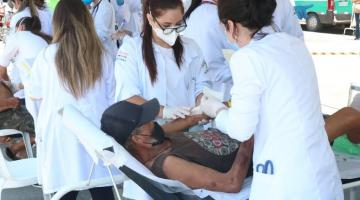 mulheres usando avental branco fazem curativo no braço de homem deitado em maca. #paratodosverem