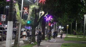 jardim da orla com árvores decoradas por luzes de natal. À direita passa a ciclovia, com bicicletas circulando. É noite. #paratodosverem 