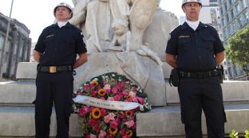 guardas municipais estão em pé diante de monumento a Braz Cubas e ao lado de coroa de flores. #paratodoverem