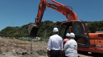 escavadeira opera em terreno. Três pessoas estão de costas para a foto usando capacete. #paratodosverem