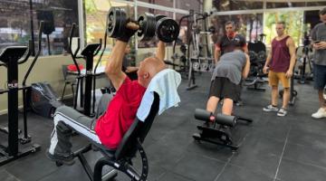 homem está sentado em banco levantando pesos com os dois braços. Ao fundo, um jovem faz exercício num equipamento para lombar. #paratodosverem 