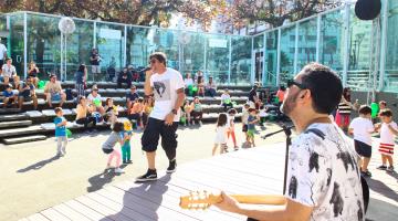 Com cerca de 20 crianças ao redor, homem canta enquanto outro toca guiterra no palco da Concha Acústica