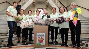 voluntárias seguram cobertores atrás de uma caixa de papelão com um selo onde se lê campanha do agasalho 