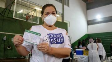 Mulher mostra carteira de vacinação, com enfermeiros ao fundo. #paratodosverem