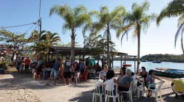 Cerca de 50 pessoas em pé e sentadas junto a mesas comem e bebem em área aberta com palmeiras junto ao mar #pracegover
