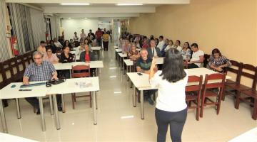 mulher de costas para a foto está falando a pessoas sentadas em uma sala de reuniões. #paratodosverem 