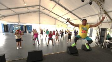 professor de dança dá salto no ar sobre o palco. Ao fundo, mulheres se movimentam. #paratodosverem 