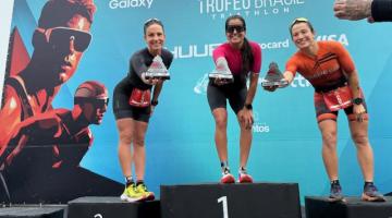três mulheres no pódio seguram seus troféus e os apontam para a foto sorrindo. #paratodosverem 
