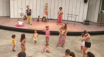 mulheres cantam e tocam em palco ao fundo. À frente, no chão, crianças brincam. #paratodosverem 