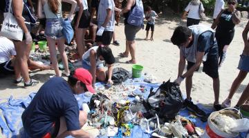 pessoas selecionam lixo coletado e separado sobre areia. #paratodosverem