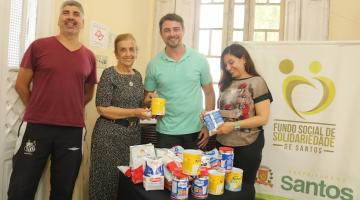 quatro pessoas em pé em torno de uma mesa onde estão latas e pacotes de leite em pó. Ao fundo, um banner do fundo social. #paratodosverem