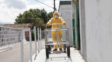 Funcionário vestindo macacão, luvas, máscara, gorro e botas empurra carrinho funerário vazio. #Paratodosverem