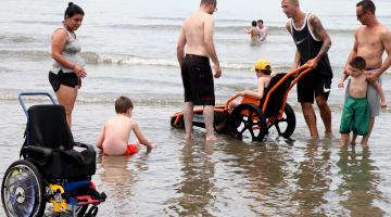 Sobre o mar raso, homem maneja cadeira anfíbia na qual adolescente está sentado com traje de banho sob observação de outras cinco pessoas