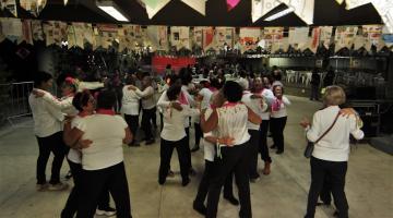 duplas de mulheres estão dançando em coreografia. Ambiente é decorado com bandeirinhas de  São João. #paratodosverem 