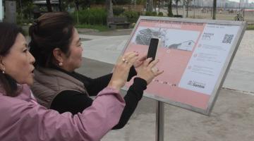 duas mulherres apontam celular para totem onde há qr code. #paratodosverem