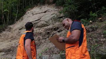 dois agentes da defesa civil estão na parte de baixo de um morro e à frente de uma encosta. Um deles faz anotações em uma prancheta. #paratodosverem