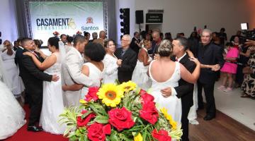casais dançam em salão. As mulheres usam vestido de noiva e os homens, terno e gravata. Em primeiro plano há um arranjo de flores. #paratodosverem 