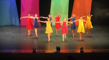 bailarinas com vestidos iguais e cores diversas apresentam coreografia sobre o palco. todas estão em pé com os braços abertos ao lado do corpo e na ponta dos pés. #paratodosverem 