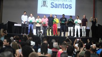 salão lotado de pessoas com palco ao fundo onde estão diversos atletas com seus troféus. #paratodosverem 