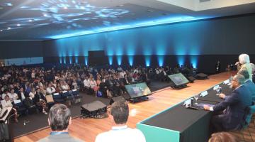palco com mesa e pessoas no auditório lotado ao fundo. #paratodosverem 