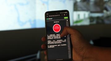 mão segura celular com botão de alerta vermelho na tela. Ao fundo, desfocado, a um mapa em um telão. #paratodosverem