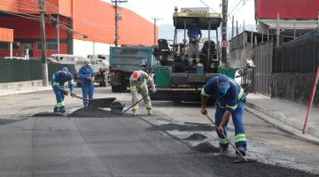 Trabalhadores de uniforme azul com enchadas na mão espalhando piche pela rua no trabalho de recapeamento