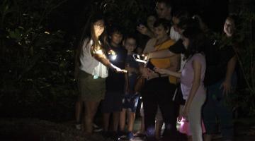 Grupo de crianças e adultos reunidos observando algo que está na palma da mão da educadora. É noite. Lanternas e luzes de celulares estão acesas. #paratodosverem 
