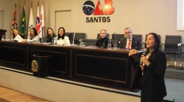 mulher fala ao microfone. Atrás dela ha um palco com seis pessoas sentadas diante de um balcão. Ao fundo, o símbolo da OAB. #paratodosverem 