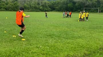 criança dá saltos em gramado em treino de futebol. Ao fundo há outras crianças e uma trave de gol mais ao fundo. #paratodosverem 